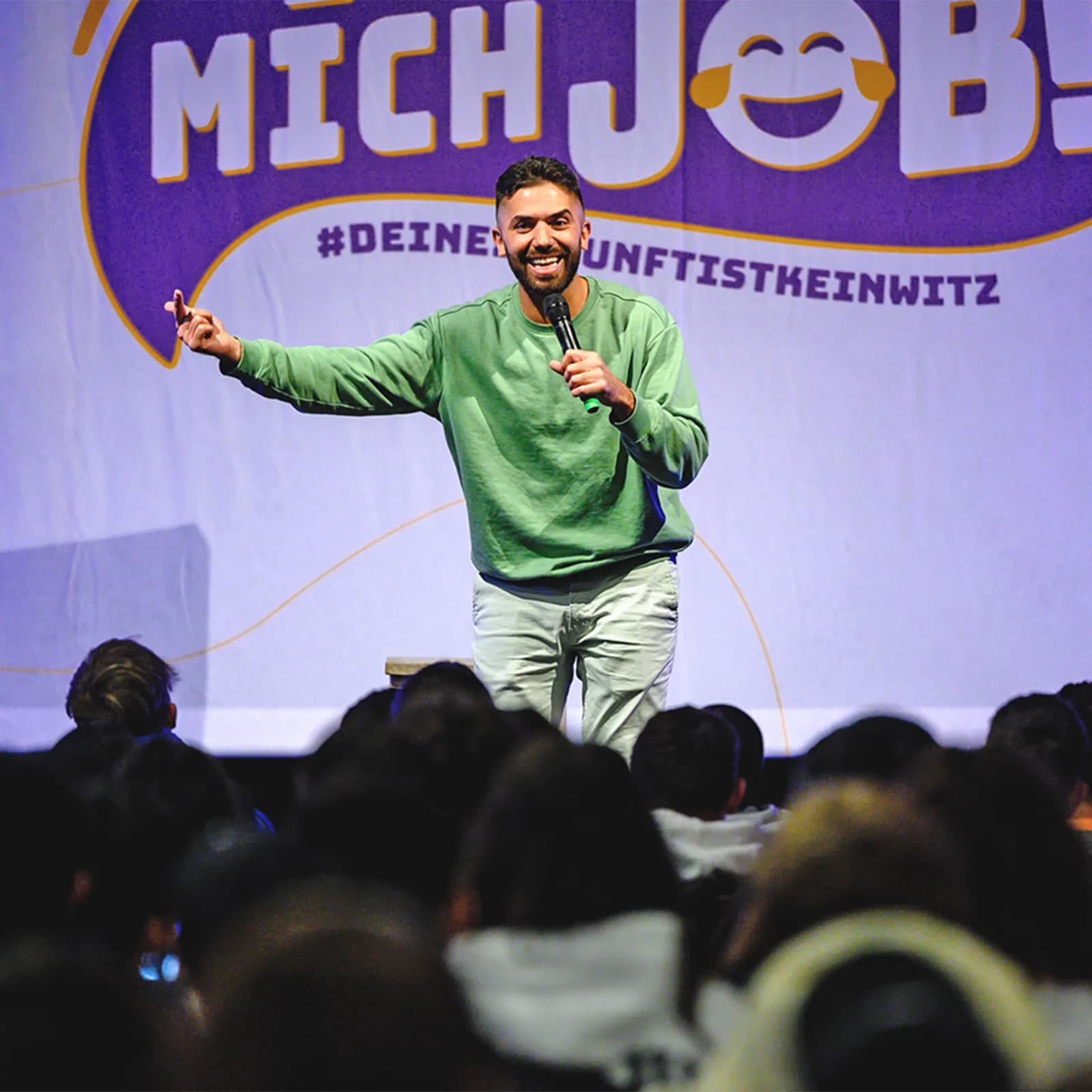 Ein Comedian steht auf der Bühne der Veranstaltung „Ich lach mich Job“ und interagiert lachend mit dem Publikum.