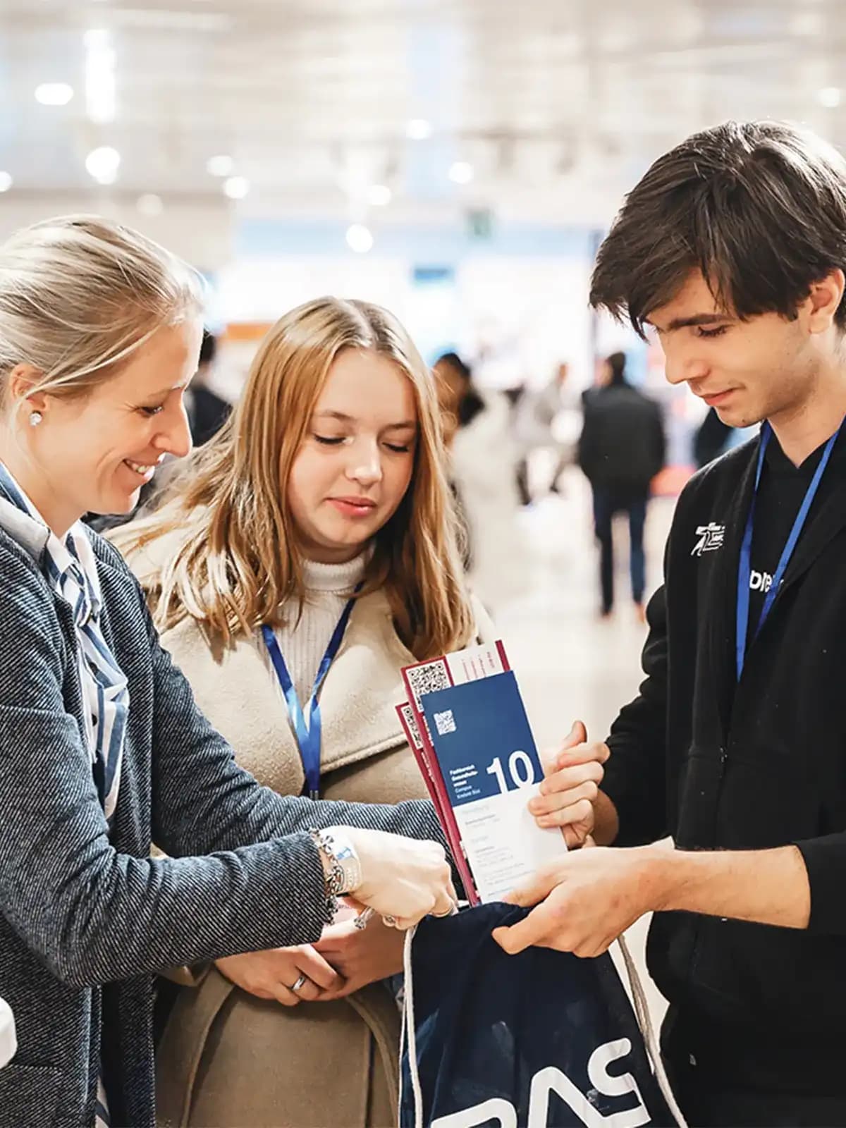 -Zwei Schülerinnen im Austausch mit einem Aussteller am Messestand, dabei werden Flyer und Infos überreicht.
