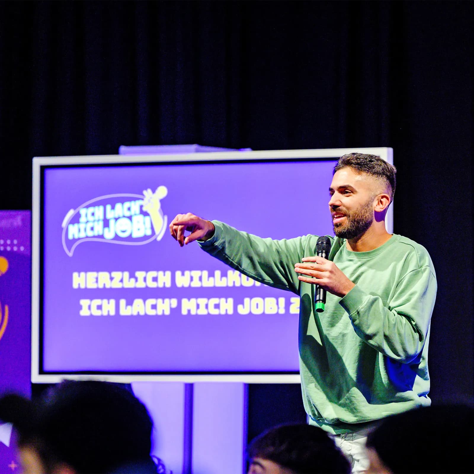 Blick in den Saal: Ein Comedian tritt bei „Ich lach mich Job“ auf, während zahlreiche Jugendliche im Publikum sitzen und aufmerksam zuhören.