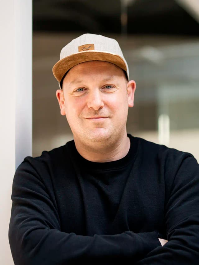 Portrait von Oliver Leonards, Geschäftsführer bei VLIP mit Schwerpunkt Konzeption & Placemaking, mit schwarzem Pullover und Cap vor modernem Bürohintergrund.